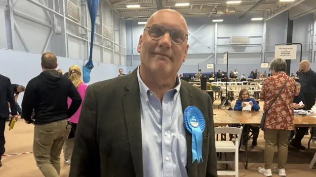 Nick Adams-King, a bald man in a suit with a blue rosette on his chest, stood in a leisure centre