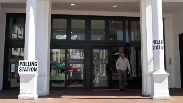 A man walking out from a large set of black doors in the front of a white building. There are two white pillars either side of the entrance with signs attached, reading 'POLLING STATION' in black lettering