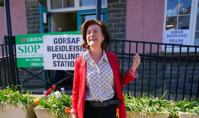 Eluned Morgan wearing a red blazer, a white patterned top while standing in front of a polling station sign