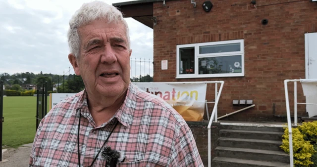 A head and shoulder shot of a man with a picnic patterned shirt and grey hair. He's standing in front of a building on the left and a cricket ground on the right.