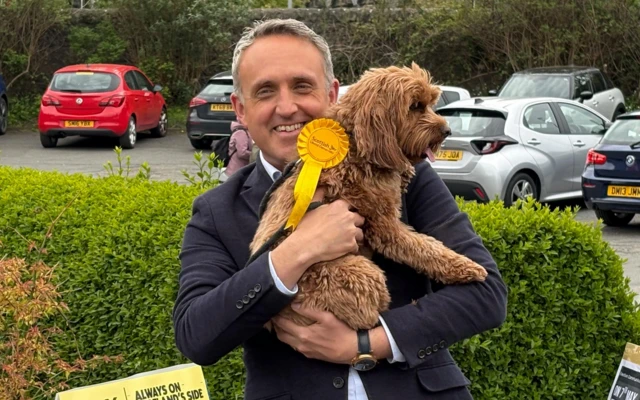 Cole Hamilton holding a small dog with a yellow rosette outdoors near a parking area and greenery.