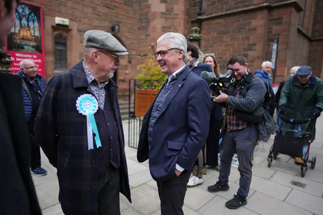 Offord talking to a man wearing a rosette, with other people and a cameraman in the background.