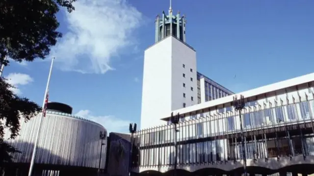 The Newcastle Civic Centre building. It is a long white building on the left hand side with a large white tower at the right end of it leading to a green tower at the top and a flagpole. On the left hand side is a round white building