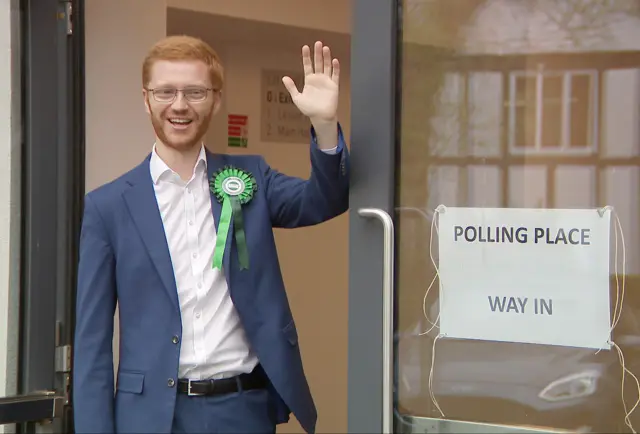 Greer wearing a green rosette waving while standing in a doorway next to a sign reading "Polling Place Way In".