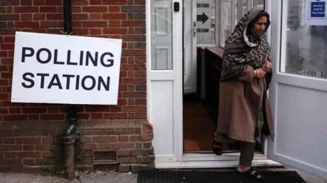 A woman in a brown dress, sandals and headscarf walks out of a red brick building that has a sign out the front saying 'polling station'