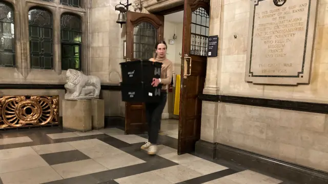 A woman walking with two ballot boxes in her hands into a town hall