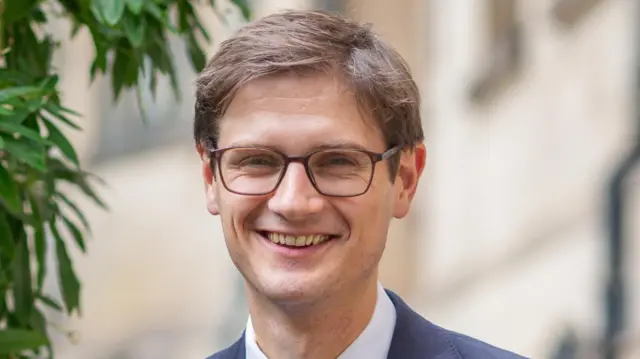 Sheffield Council leader Tom Hunt stands smiling. He is wearing a suit and glasses.