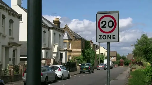 A street of two-storey houses, a mixture of detached and terraced housing. There is a road passing by, on which several cars are parked. In the foregound is a 20mph zone sign. There are trees to the right.
