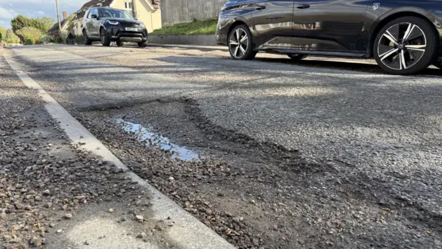 A pothole in a road with gravel spreading out from it and cars passing in the background.