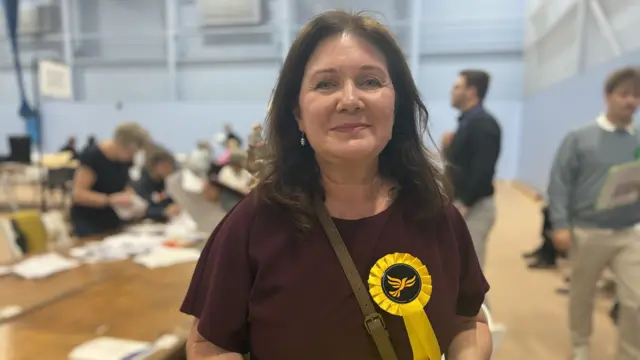 Sally Yalden, a woman in a maroon top stood in a leisure centre with a yellow rosette on her chest.