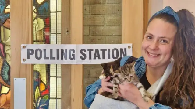 Leigh wears a carrier with a number of kittens inside. She stands in front of a sign reading 'polling station'