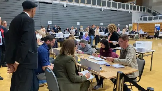 Lots of people sit in a hall at foldable tables, counting votes. It's a busy atmosphere