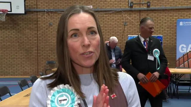 Fiona Pearce is speaking to an interviewer behind the camera and has her hands clapped together. She has long brown hair and is wearing a white a turquoise rosette which reads: "Reform UK."