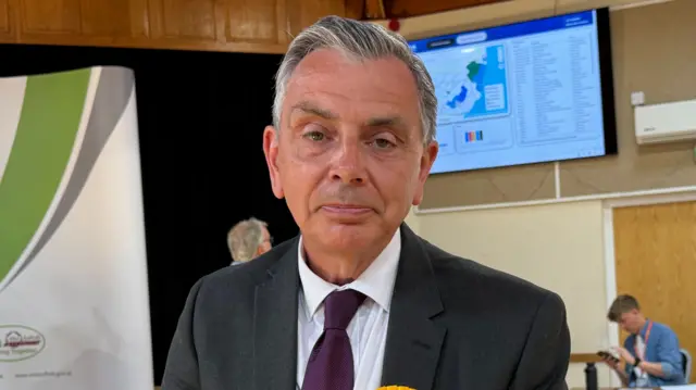 A man with grey hair looks at the camera. He wears a black suit jacket with a white shirt and dark purple tie.