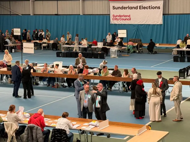 A view of the election count in Sunderland at Silksworth Lesiure Centre. On the far wall is a white banner with the words Sunderland Elections and Sunderland City Council. There are tables with rows of vote counters and candidates standing in between them ro