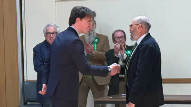 Two men wearing suits shake hands as they smile at each other. One is younger than the other.
