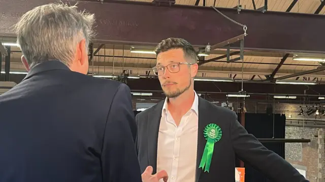 Tom Butcher is doing a TV interview - he is facing the interviewer wearing a suit jacket and white shirt with a green rosette attached.