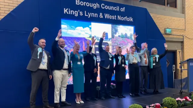 A group of men and women stand in a line on a stage with their hands in the air celebrating victory for their political party.