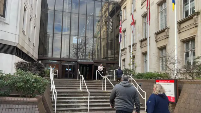 A large building with light-coloured walls, steps leading up to it and a glass frontage. Flagpoles also line the route into the building.