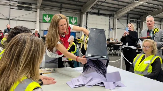 a woman pour a ballot vox of purple voting slips onto a desk. There are several people in hivis vests sitting around the desk.
