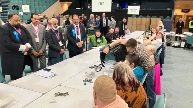 A large table goes across the screen. One person is holding a bucket full of ballot papers and is pouring them on to the table. Several people are watching from the left side of the table as people are sitting on chairs on the other side.