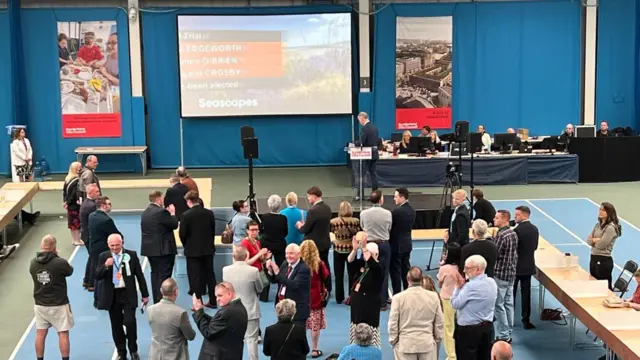 Dozens of people stand around the sports hall looking up at the small black stage which has been set up. The walls behind are draped in blue curtains and there are red Sunderland city banners up as well. Plus a projector screen showing some candidates' names.
