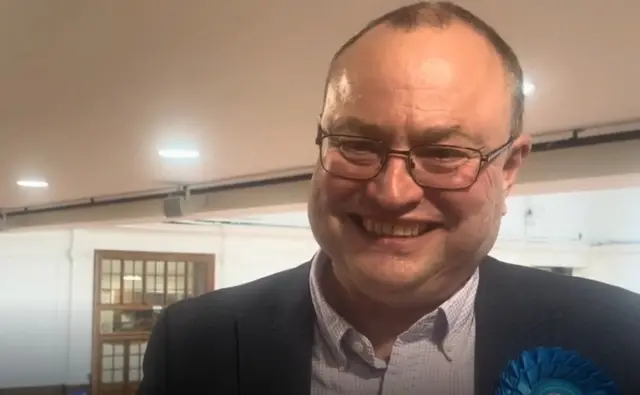A smiling middle aged man in a suit and wearing a blue rosette being interviewed