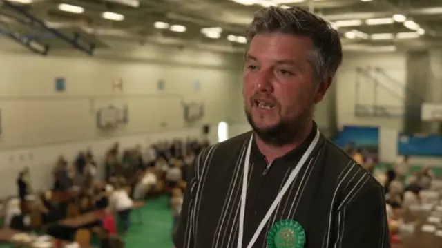 Man in grey shirt wearing green rosette giving tv interview in large hall with people behind him
