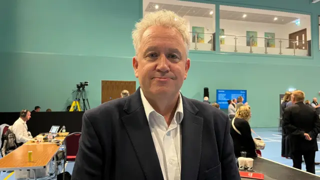 A man in a dark suit and white shirt looks at the camera while at an election count