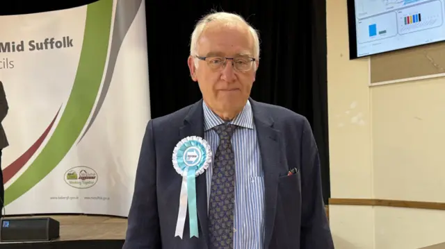 A man wearing a navy blue suit, blue and white striped shirt and a tie looks at the camera. He has a light blue rosette pinned to his chest. He has grey hair and wears glasses.