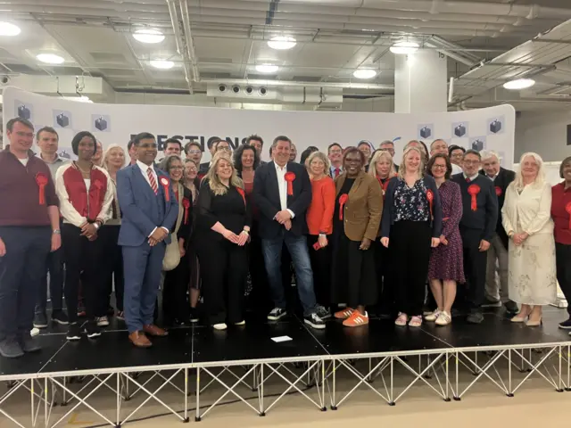 A group of Labour councillors, all wearing red rosettes, stand on a stage posing for a photo