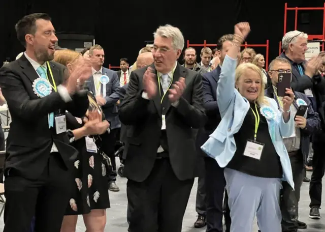 Men and women in sky blue rosettes and suit jackets cheer and clap in a large hall with people behind them.