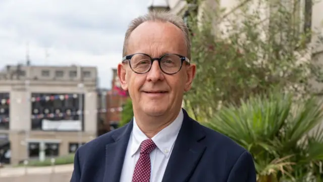 A man with round glasses in a suit stands infront of a blurred background of a town council