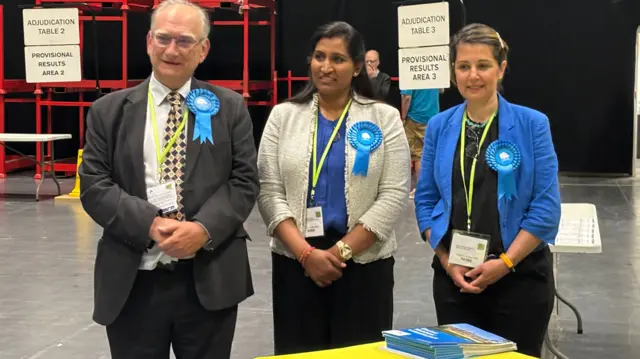 A man in a grey suit, woman in blue top and white cardigan and woman in blue jacket are all wearing blue rosettes and standing behind a table at an election count.
