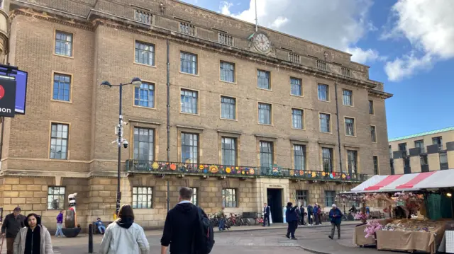 A four-storey Victorian style building with a balcony above the first floor. There is a clock at the top. People are walking on the pavement in front.