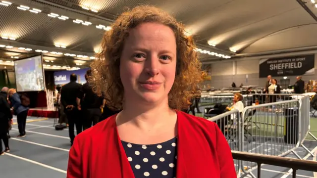 Olivia Blake MP looks at the camera. She has curly red hair and is wearing a red jacket over a black top with white spots.
