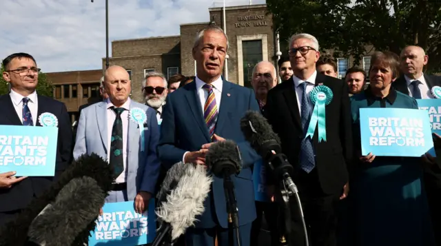 Nigel Farage surrounded by other people holding Reform banners