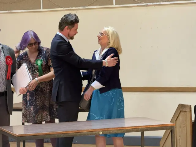 A man and woman stand close together in joy. The man has his hands resting on the woman's arms as they look at each other. He wears a black suit and has dark hair. The woman has short blonde hair and wears a black cardigan with a blue skirt and a white shirt.