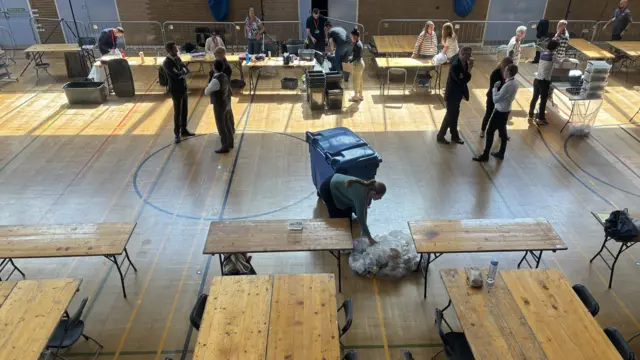 A sports hall where people are cleaning up after an election count. Some people can be seen standing and talking in some groups around the hall