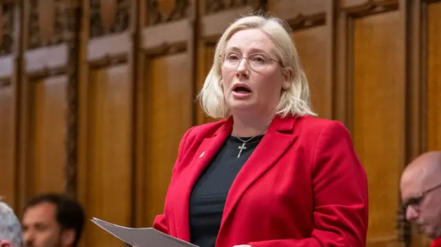 Emma Lewell is standing and speaking in the House of Commons. She has mid-length blonde hair and glasses. She is wearing a black shirt and a red blazer.
