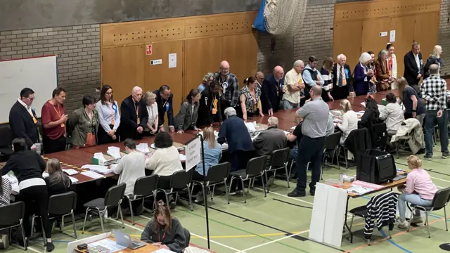 There is a large brown table which goes across a room. People are standing on one side of the table and on the other side people are sitting on chairs counting ballot papers.