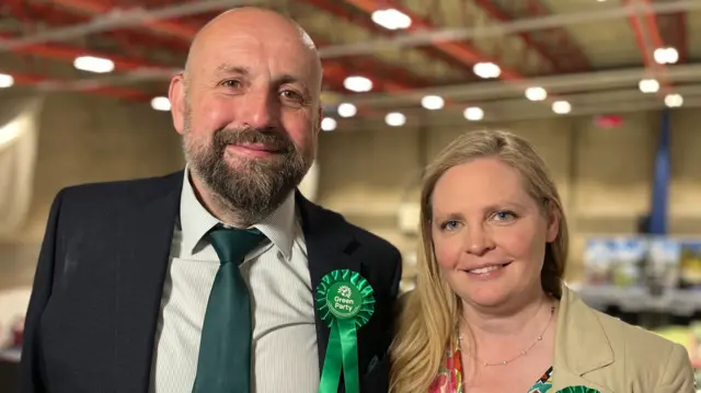 Stephen Ferguson and Lara Davenport-Ray are standing side by side. Ferguson, who is on the left, has a short brown and grey beard. He is wearing a cream shirt, green tie and black suit jacket. He is wearing a Green Party rosette. Davenport-Ray is on the right. She has long blonde hair and is wearing a floral dress and beige blazer.
