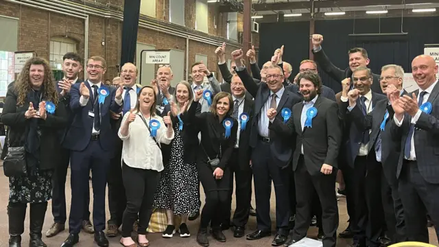 A group of Conservative candidates celebrating their win, posing for a picture wearing blue rosettes.