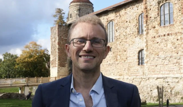Lee Scordis wearing light shirt and dark blazer. He has black rimmed glasses and light, fluffy close-cropped hair