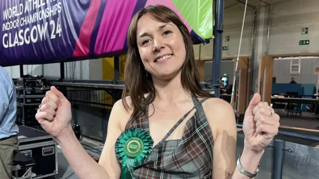 Holly Bruce has her fists in the air, celebrating. She is wearing a green tartan dress and a Scottish Greens rosette