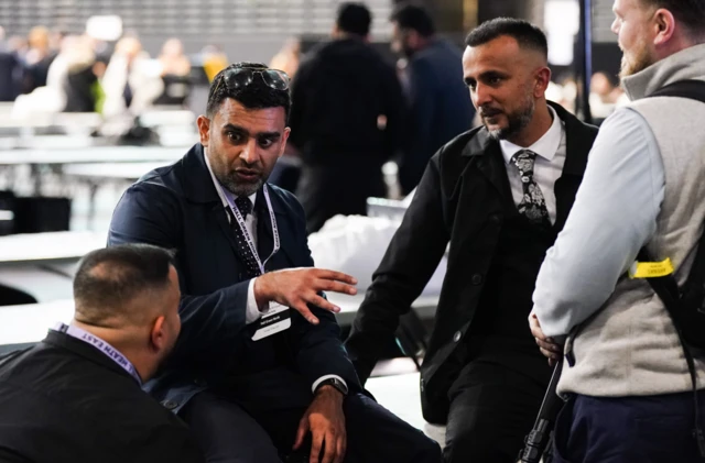 A group of four men gather around and talk. Three of them wear suits and ties.
