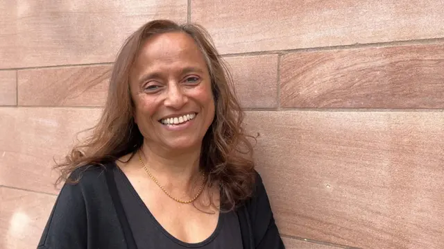 Malkit has light brown hair and is wearing a black top with a gold necklace. She is smiling and standing in front of a red sandstone wall.