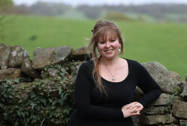 Delyth Jewell stands in front of a stone wall by a field smiling at the camera.