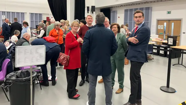 Eluned Morgan wearing a green suit surrounded by members of her team. There are a number of people behind them counting votes.