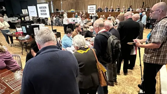 People stood over tables in a large town hall.